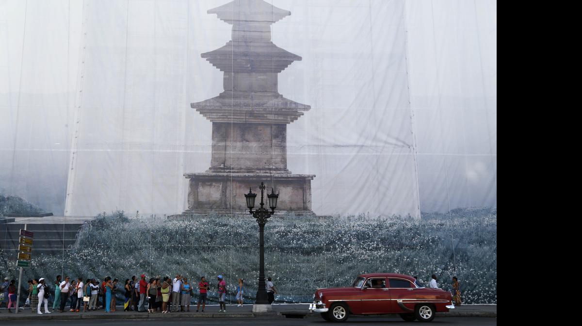 
Pasajeros esperan un autobús en La Habana. Al fondo una fotografía del artista coreano Han Sungpil desplegada en la fachada de un edificio, una de las obras que participal en la bienal.
