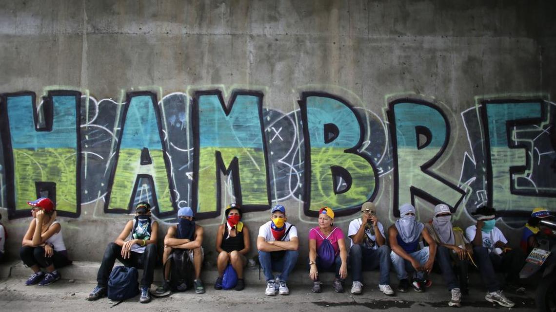 Manifestantes contra el régimen de Nicolás Maduro se sientan en una acera y detrás hay una pared con la palabra “hambre”, el 1 de julio de 2017.