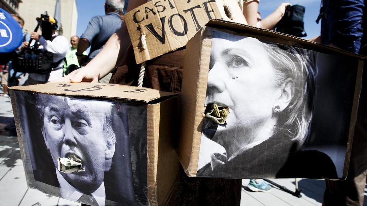 Ira Birch, una manifestante de Richmond, Virginia, sostiene dos fotografías de Donald Trump (i) y de Hillary Clinton (d) con billetes de dólares en su boca durante una manifestación llamada ‘Wall Off Trump’ (Amurrallar a Trump) en Cleveland, Ohio, el 20 de julio de 2016.
