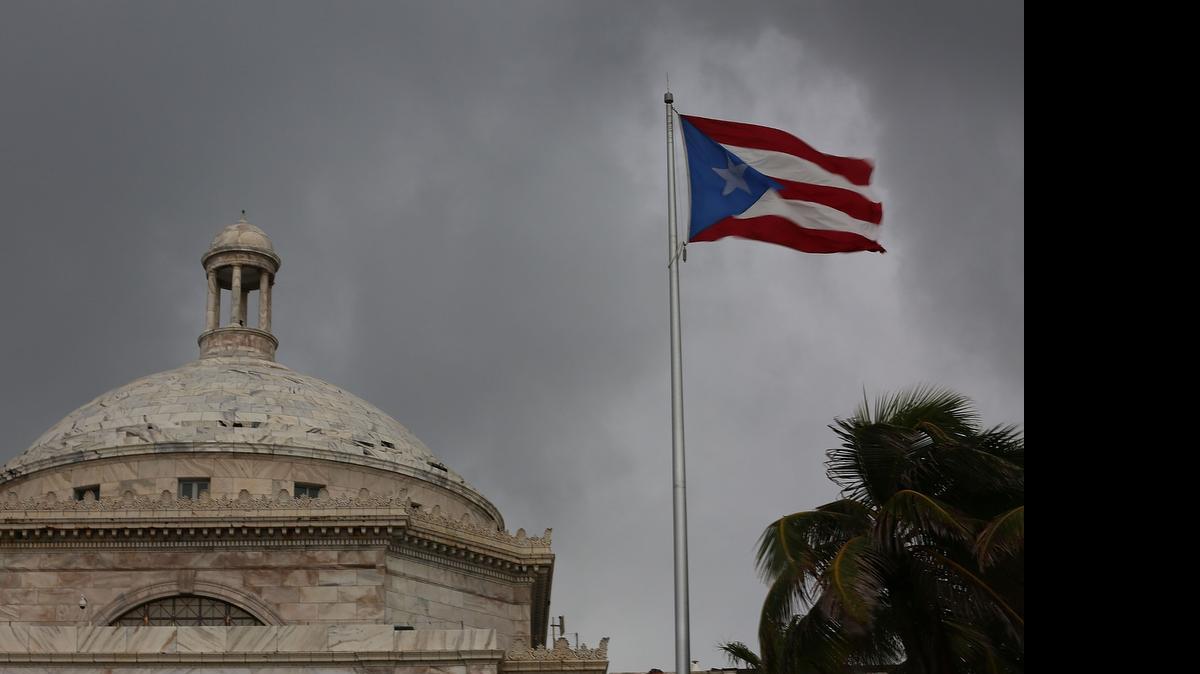 
El Capitolio y la bandera puertorriqueña en un cielo gris.


