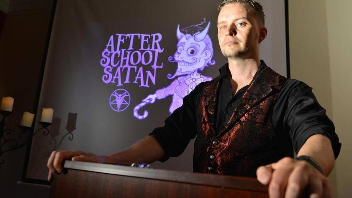 Doug Mesner, también conocido como “Lucien Greaves”, posa junto al logo del programa de actividades extracurriculares “After School Satan Club”.