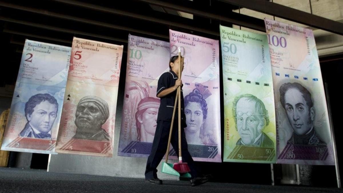 Una mujer pasa con implementos de limpieza frente a afiches de billetes venezolanos en la sede del Banco Centra de Venezuala, el año pasado, Caracas.