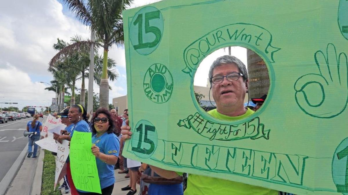 Jaime Ordoñez (d) muestra una pancarta con un billete de 15 dólares frente al Walmart de la Calle 49 y la Avenida 4 del Oeste, en Hialeah.
