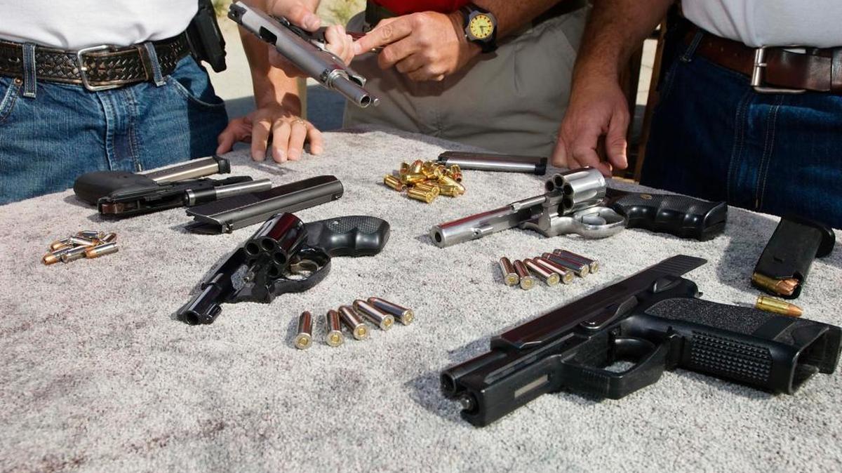 Tres hombres observan armas y municiones en una mesa.