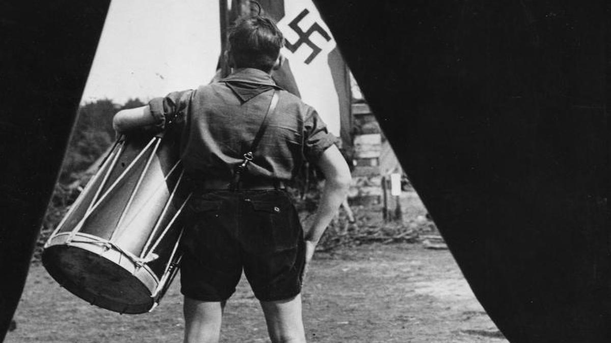 
Un adolescente miembro de la Juventud Hitleriana (’Hitlerjugend’), uniformado y portando un tambor, observa una bandera nazi, en la década de 1930 en Alemania, cuando la juventud alemana fue indoctrinada contra los judíos. 
