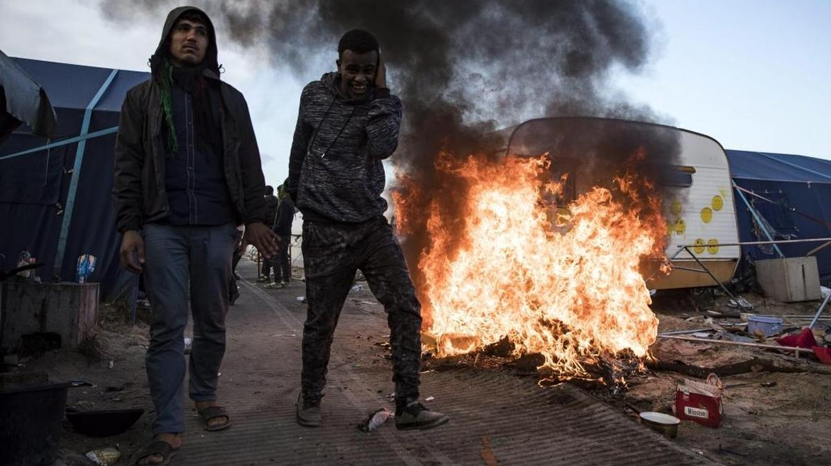 Dos inmigrantes caminan junto a una estructura en llamas durante el proceso de desmantelamiento de algunas de las tiendas que han configurado el campamento de inmigrantes de Calais, conocido como “la Jungla”.