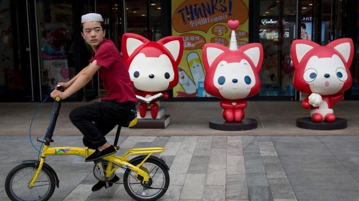 Un hombre  con un casquillo de minoría étnica pasa con su bicicleta frente a  unas mascotas de dibujos animados afuera de un centro comercial en Beijing, China, el miércoles, 17 de agosto de 2016. 
Según la Organización Mundial de la Propiedad Intelectual de las Naciones Unidas China se ha sumado a la lista de las  25 mayores economías de innovación, en el mundo, por primera vez.