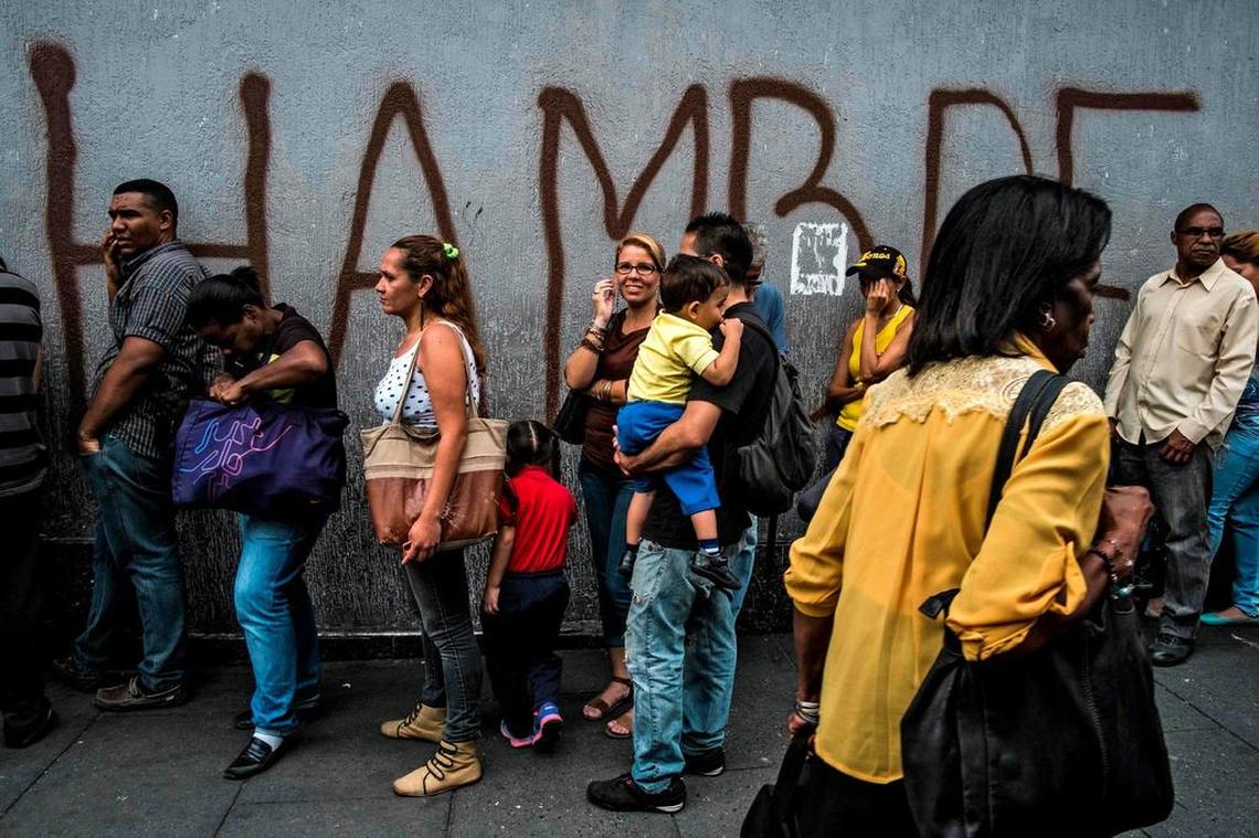 People wait next to a wall with graffiti reading “Hunger” in Caracas on July 23, 2018.  The financial and humanitarian crisis in Venezuela has resulted in a mass exodus from the country.
