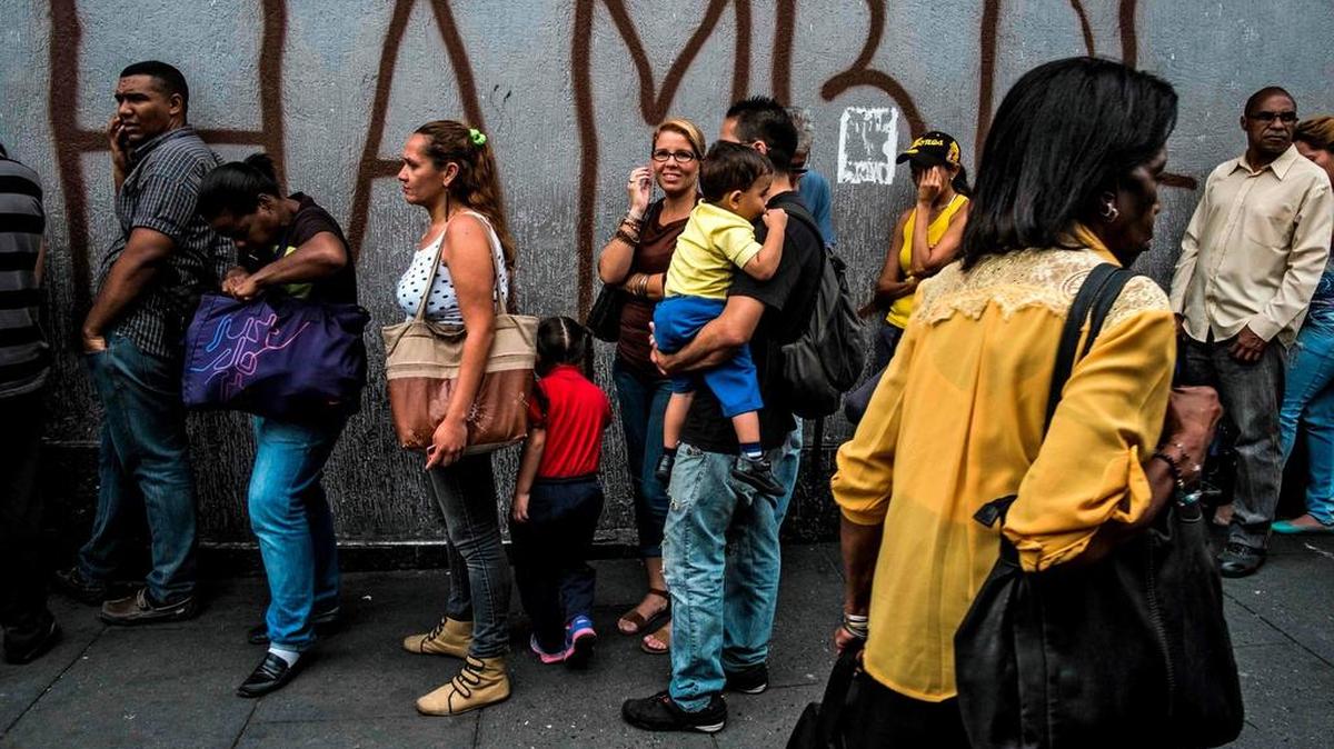 People wait next to a wall with graffiti reading “Hunger” in Caracas on July 23, 2018.  The financial and humanitarian crisis in Venezuela has resulted in a mass exodus from the country.