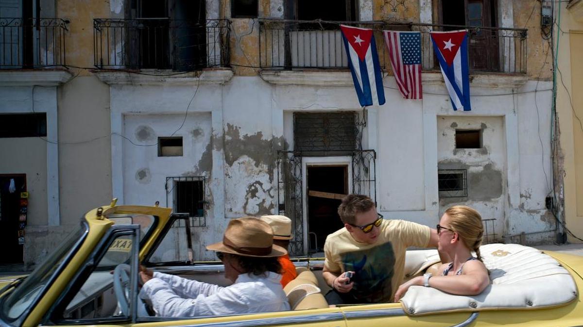 Turistas viajan en un auto americano vintage pasando frente a un balcón decorado con banderas cubanas y de Estados Unidos, en La Habana.