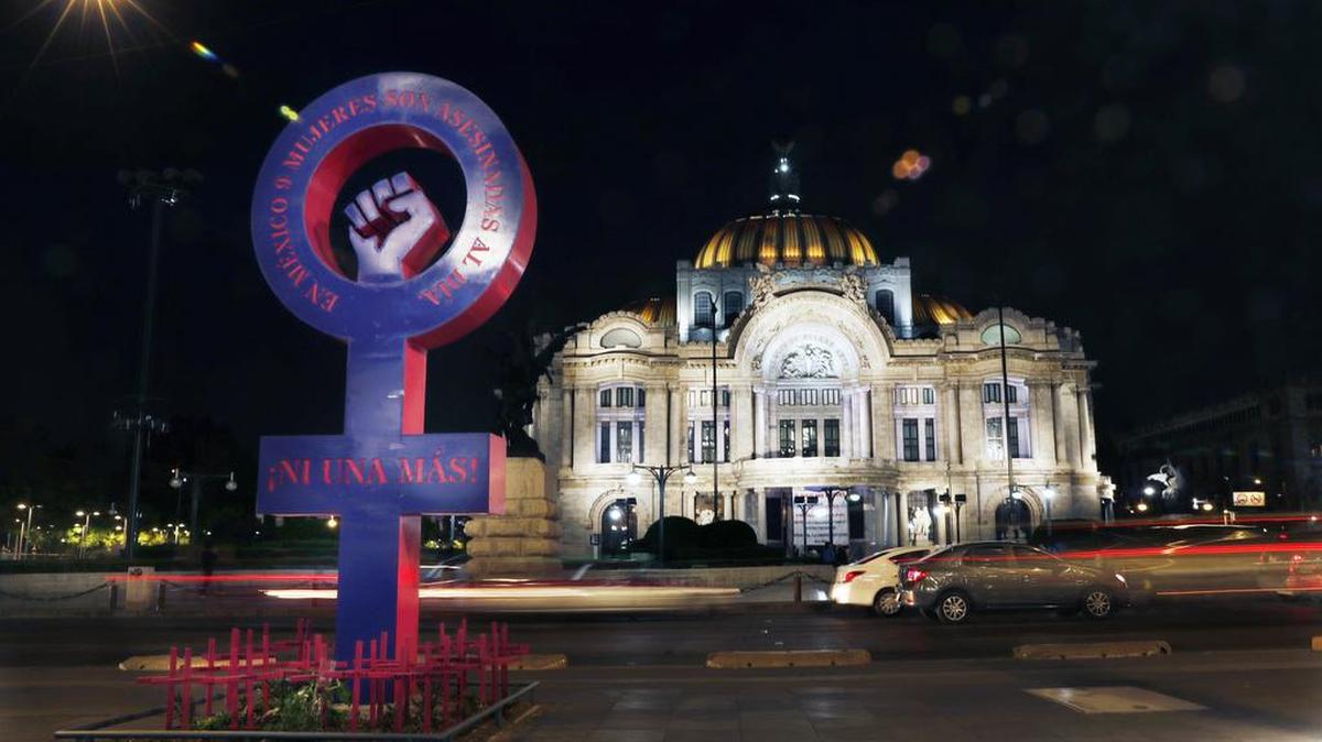 Un círculo púrpura con una cruz debajo –el símbolo reconocido internacionalmente para el género femenino– y un puño levantado y cerrado exigen “¡Ni una más!”, en relación con los feminicidios. En la base, hay un jardín lúgubre con cruces rosas que tienen inscrito el nombre de los estados del país. Localizado frente al Palacio de Bellas Artes en la Ciudad de México.