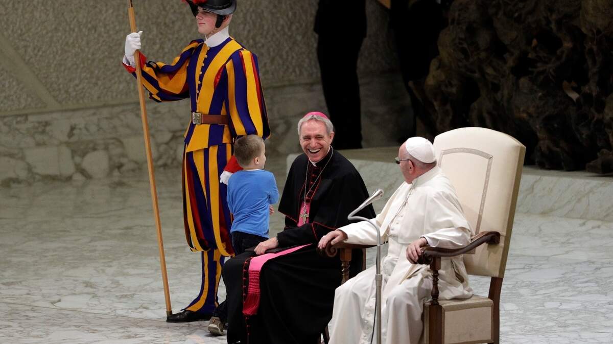 El papa Francisco y el arzobispo George Gaenswein sonríen en el momento en que un niño empieza jugar con el guardia suizo, durante la audiencia papal del 28 de noviembre de 2018 en el Salón Pablo VI del Vaticano. Después se reveló que el niño argentino es autista y no puede hablar.