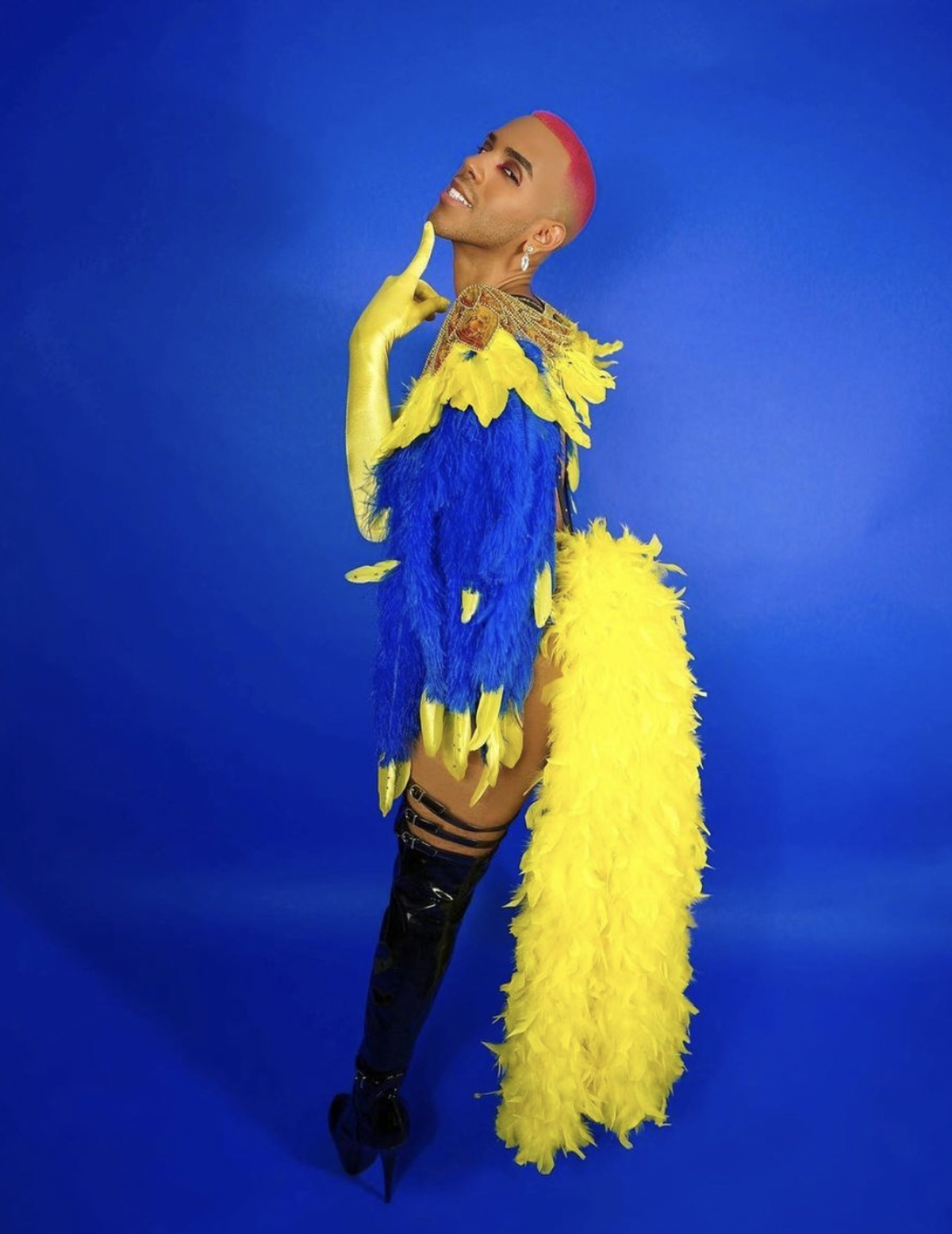 Artista Sin Silva posa delante de un fondo azul con un traje azul con plumas amarillas y botas negras.
