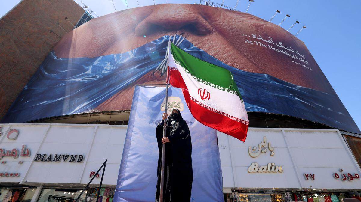 Una mujer sostiene la bandera iraní mientras permanece frente a un cartel antiestadounidense relacionado con el presidente Donald Trump y el estrecho de Ormuz, instalado en un edificio de la plaza Valiasr en Teherán el 10 de mayo de 2026.