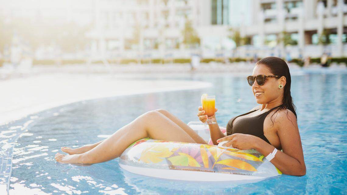 Beautiful woman in pool lay on her back in inflatable ring at sunset, she looking away and smiling, drinking summer orange cocktail.
