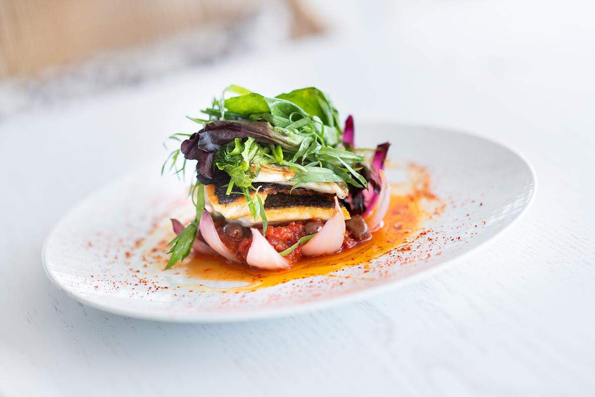 La exquisita lubina a la provençale, fileteada con piel al horno, se sirve con la típica mermelada de tomate francesa agridulce al punto, olivas y ensalada. Restaurante Juvia.