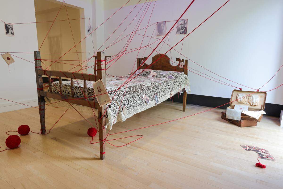 'Ancestral Lines-Inherited Ties', 2025, de Marine Fonteyne. Antigua cama de familia, cobertor con encaje de Brujas (Bélgica), fotografías familiares bordadas, lazo rojo y otros objetos personales.