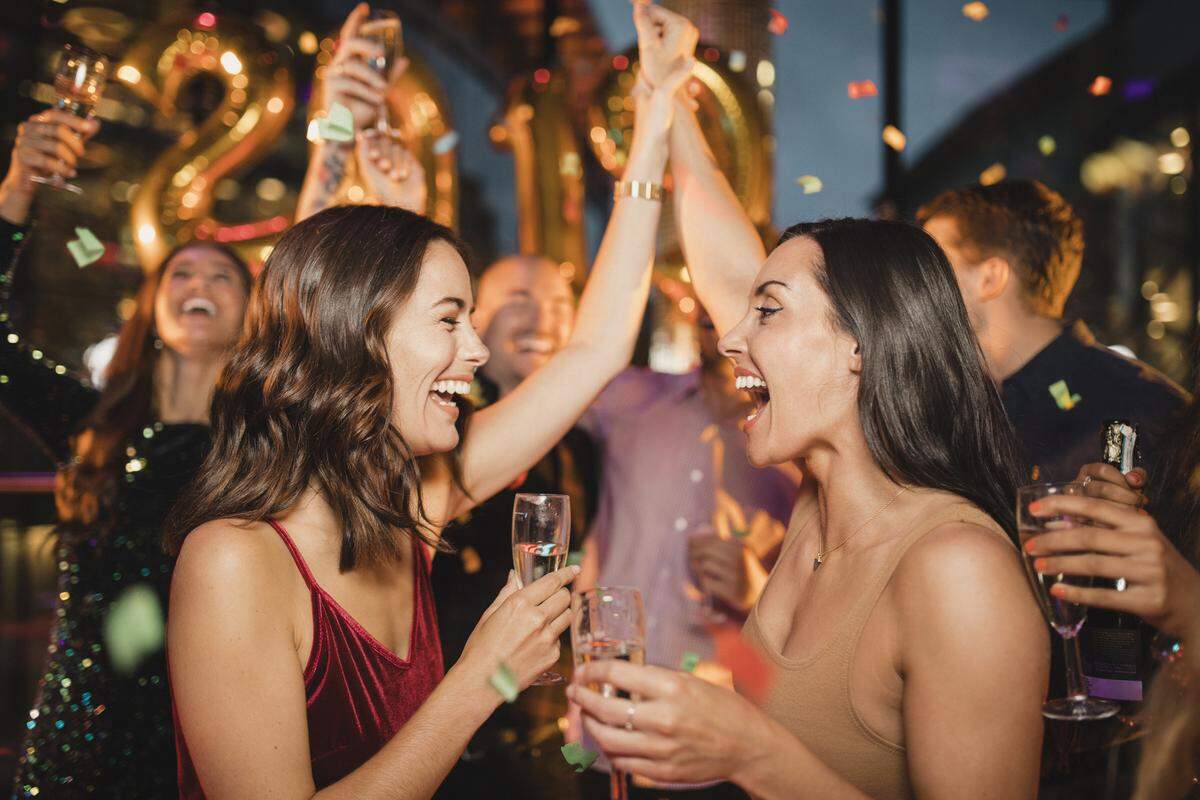 Two friends dancing among other people while celebrating the new year with a glass of champagne, balloons and confetti.