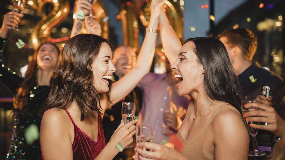 Two friends dancing among other people while celebrating the new year with a glass of champagne, balloons and confetti.