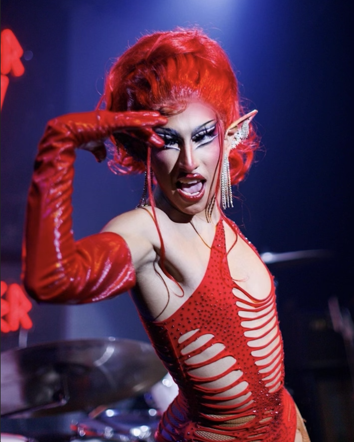Artista de drag Viola Putx se ve posando en un disfraz completamente rojo — incluyendo guantes largos y peluca. Viola tiene puesto orejas estilo duende.