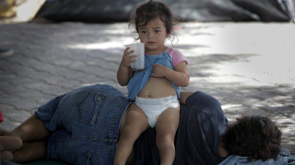 Matamoros, Mexico, August 1, 2019- A child sits on her mother under the shade of a tree in a small migrant camp right next to the Gateway International Bridge in Matamoros, Mexico. Those migrants that have some money usually rent a room to wait out the time it takes to get asylum in the United States, those who don’t usually stay in the small camp by the bridge.