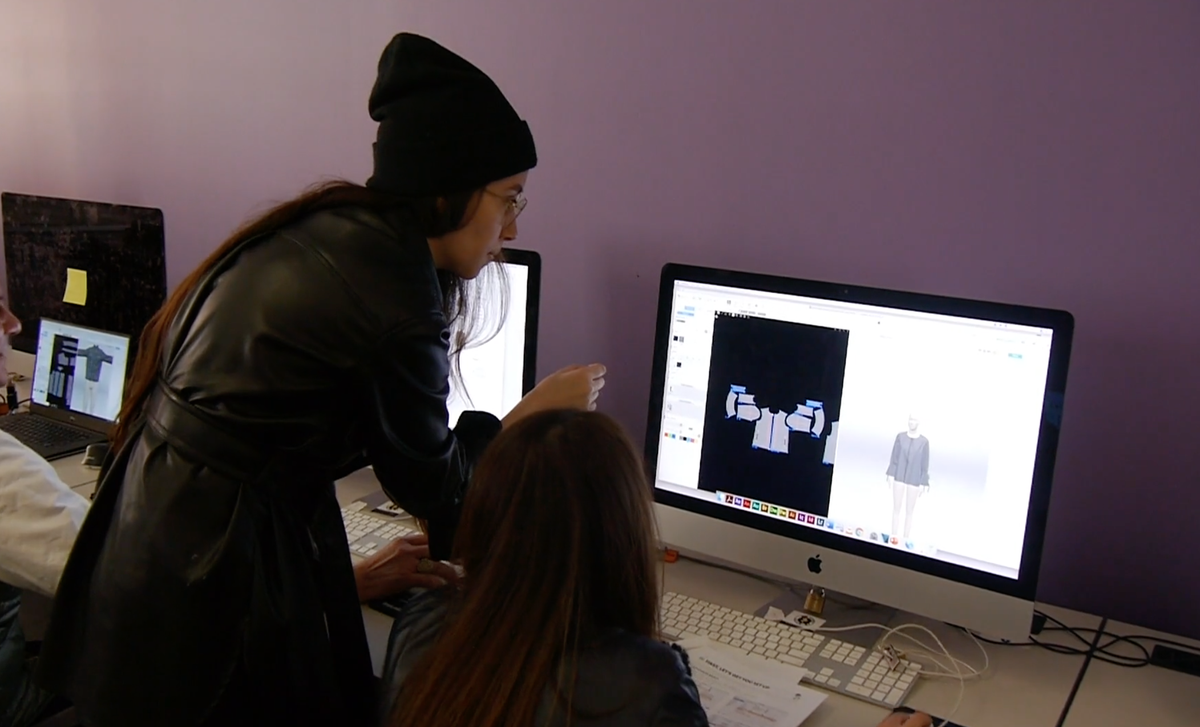 Diseñadora Karina Ochoa de Baker da una clase sobre el diseño de moda en 3D. Ayuda a una estudiante con su computadora. Están en un cuarto con paredes moradas.