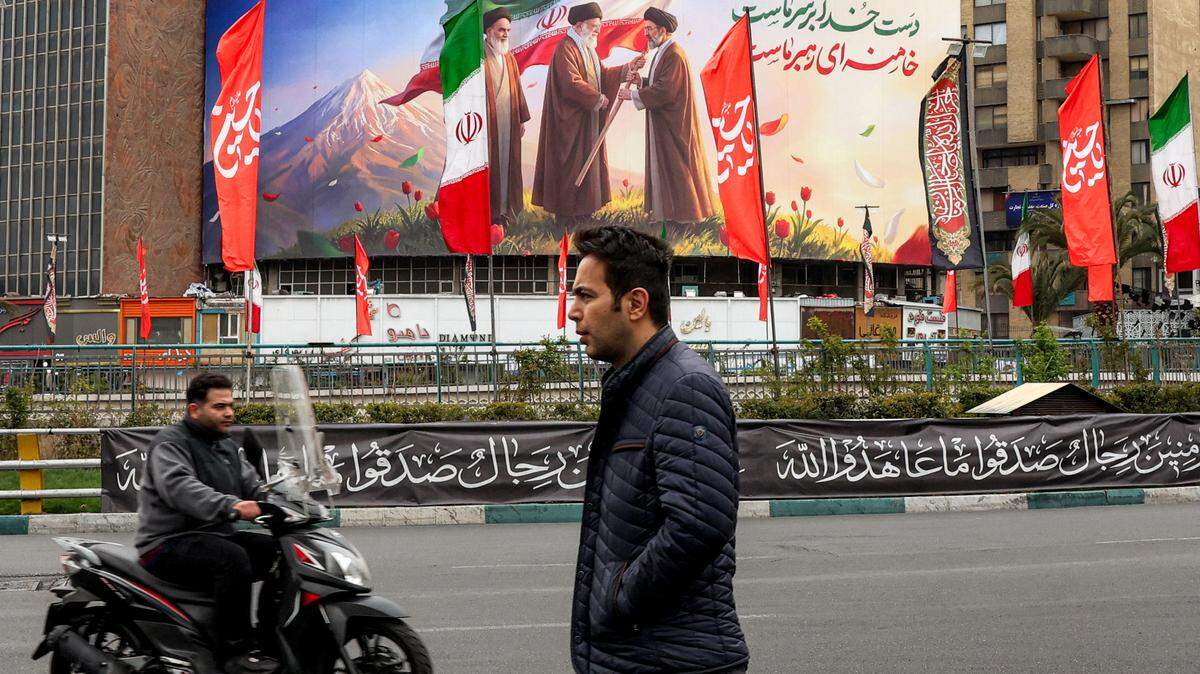 Un hombre camina junto a un motociclista cerca de un cartel exhibido en la plaza Valiasr, en el centro de Teherán, el 10 de marzo de 2026, que muestra al difunto líder supremo de Irán, el ayatolá Ruhollah Khomeini (izq.), observando cómo su sucesor, el también fallecido ayatolá Alí Jameneí (centro), entrega una bandera nacional a su hijo y nuevo líder supremo, Mojtaba Jameneí (der.).