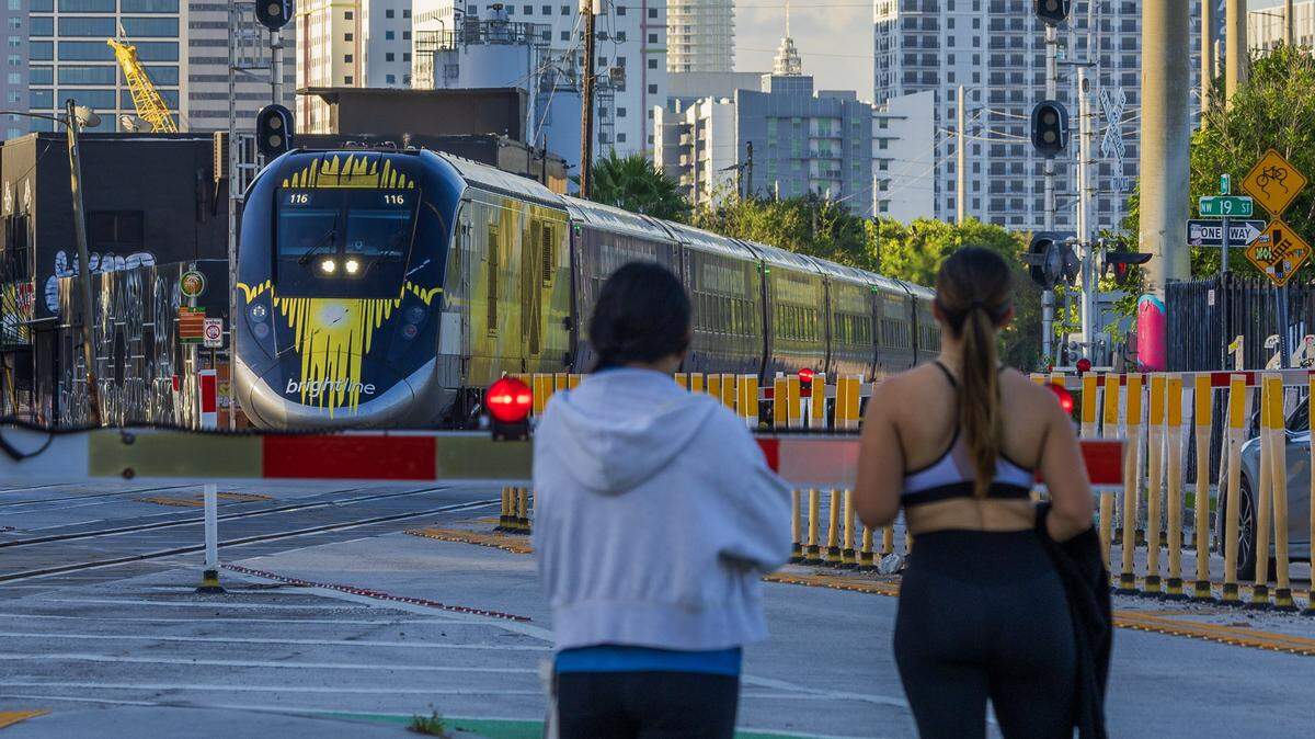 Peatones esperan para cruzar mientras un tren Brightline se dirige al norte en la intersección de la calle 20 y Miami Avenue.