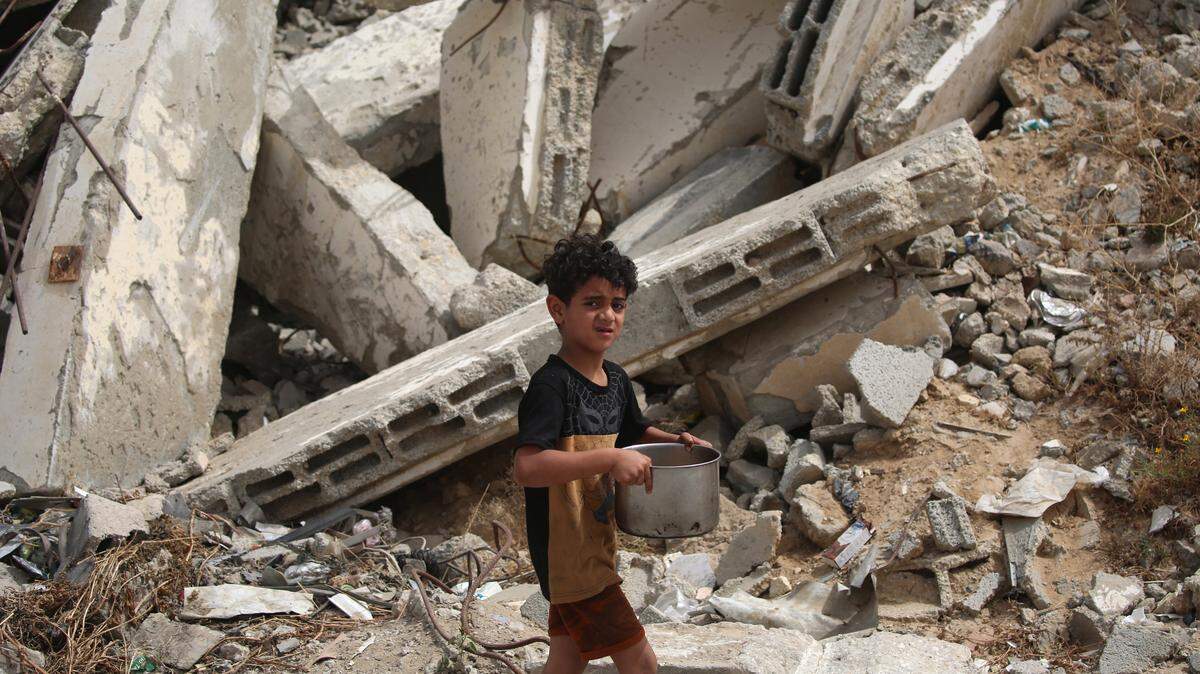 Un niño palestino desplazado lleva una olla con comida distribuida por una cocina solidaria, pasando junto a edificios destruidos por el ejército israelí, en el campo de refugiados de Bureij, en el centro de la Franja de Gaza, el 18 de mayo de 2026.
