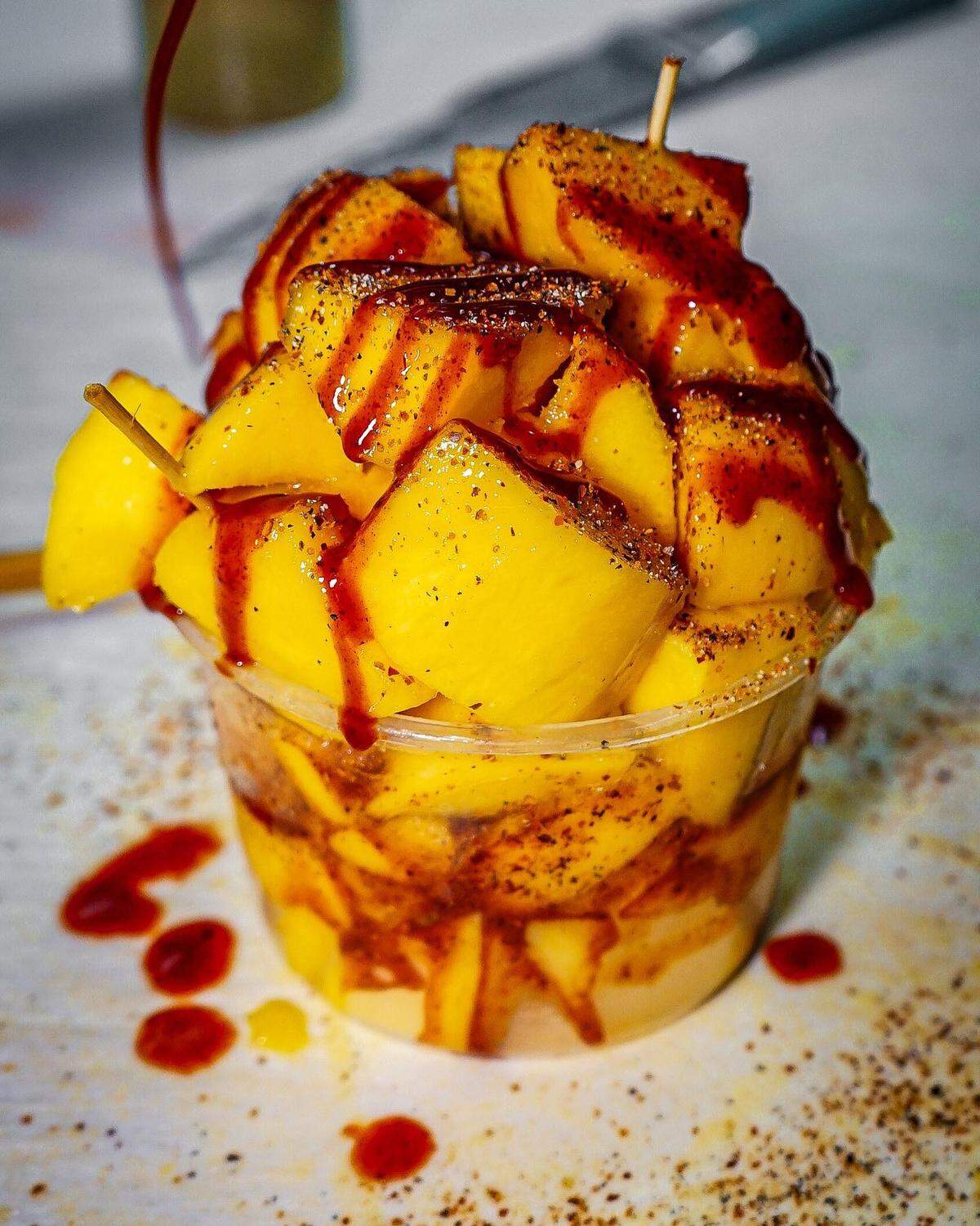 Un delicioso vaso de mango picado en trozos bañados en salsa chamoy y espolvoreados con Tajín, un aclamado condimento mexicano que les da un toque salado, cítrico y un poco picante.