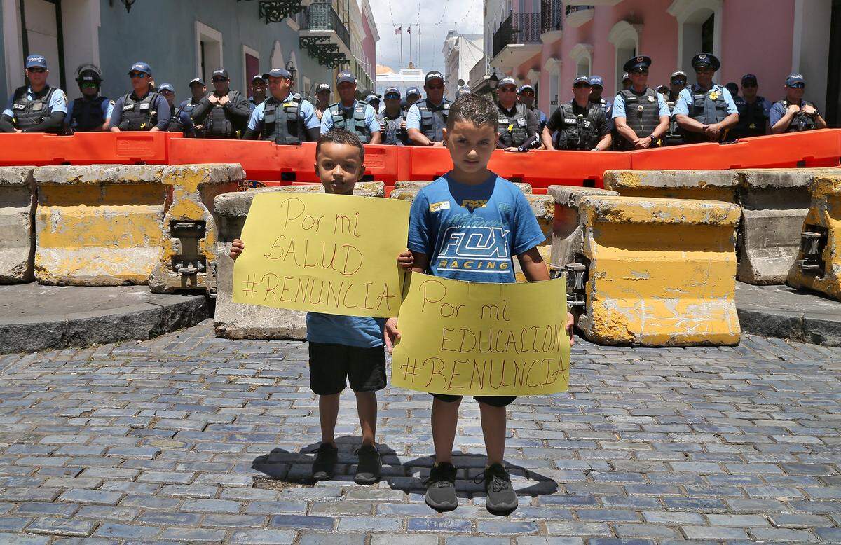 Kids Dylan (izq) y Roy Salgado, con carteles frente a un perímetro de seguridad de la policía en el Viejo San Juan, en que piden la renuncia al gobernador Ricardo Rosselló cerca de La Fortaleza, el miércoles 17 de julio de 2019.