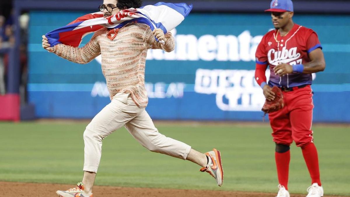 El periodista cubano Carlos Manuel Álvarez que lleva una bandera cubana con las palabras Patria y Vida corre en el campo mientras el infielder cubano Andy Ibáñez (77) observa durante el juego entre Estados Unidos y Cuba en la semifinal del Clásico Mundial de Béisbol en el LoanDepot Park en Miami, Fla. el domingo, 19 de marzo de 2023.