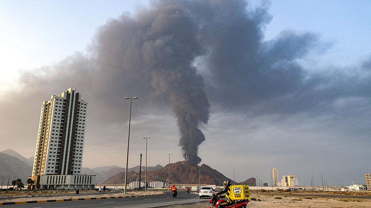Los repartidores viajan en motocicletas a lo largo de una carretera mientras detrás se eleva una alta columna de humo luego de una explosión en la zona industrial de Fujairah, en los Emiratos Árabes Unidos, el martes 3 de marzo de 2026.