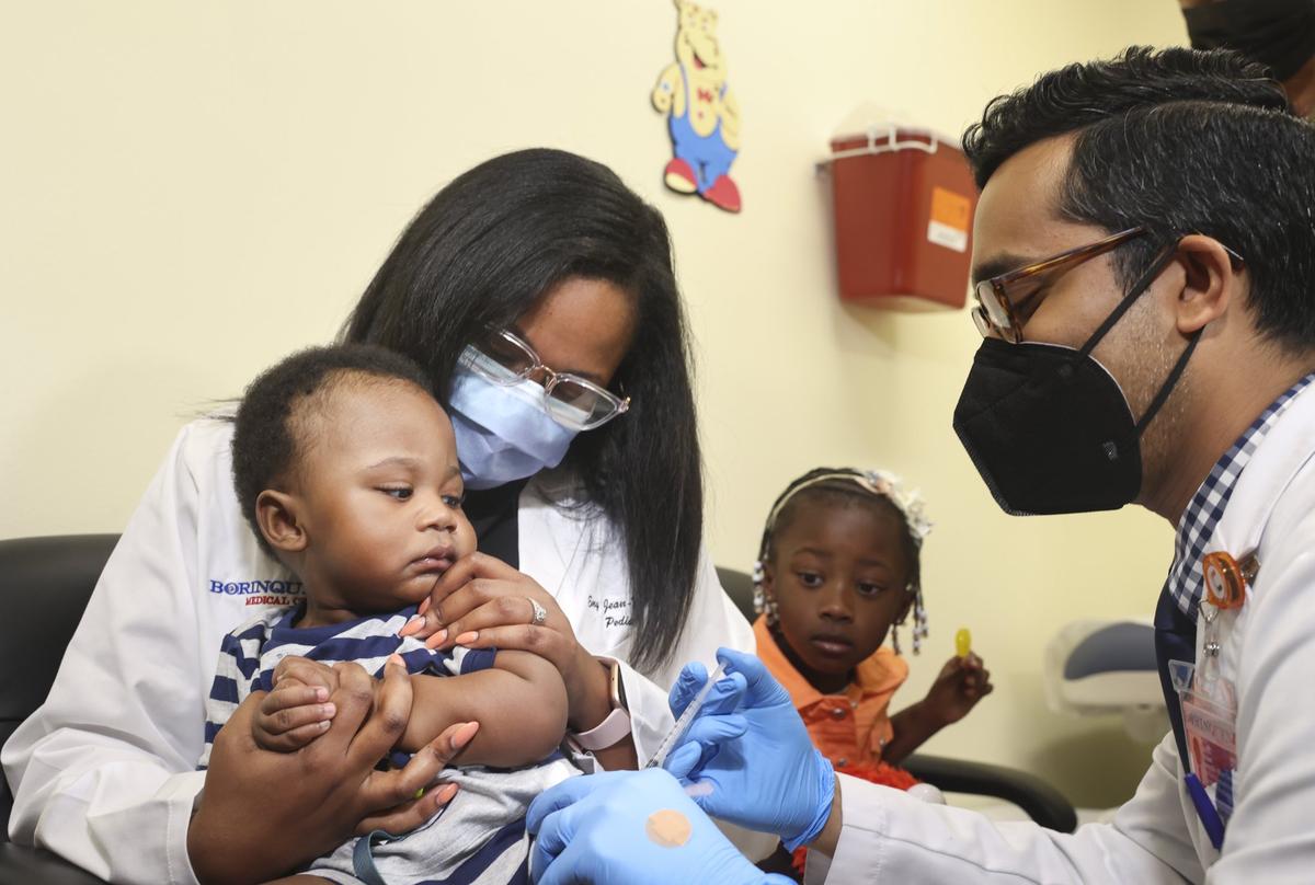 El Dr. Nizar Dowla, a la derecha, le administra la vacuna COVID-19 a Deji Adebayo, de nueve meses, mientras su madre, la Dra. Emy Jean-Marie, y su hermana, Emiola Adebayo, observan durante una campaña para niños menores de cinco años en martes 28 de junio de 2022 en Borinquen Health Care Center en Miami. La subsecretaria de Salud, la almirante Rachel Levine, y la alcaldesa de Miami-Dade, Daniella Levine Cava, asistieron a la campaña.