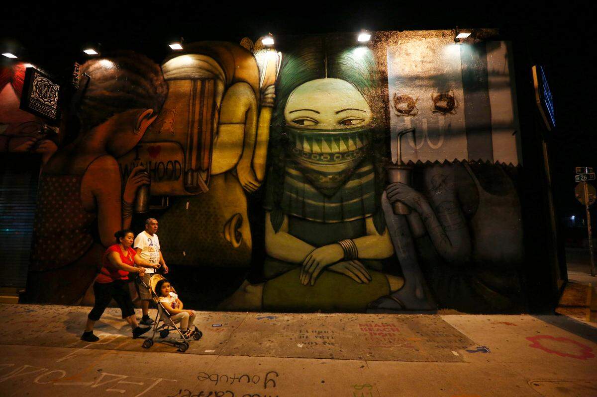 Una pareja que empuja a un niño en la carriola pasa junto a una pintura mural enmascarada en el restaurante Wynwood Kitchen & Bar. El distrito de artes de Wynwood, normalmente bullicioso, estaba desprovisto de sus multitudes habituales el miércoles por la noche, 1 de abril de 2020.