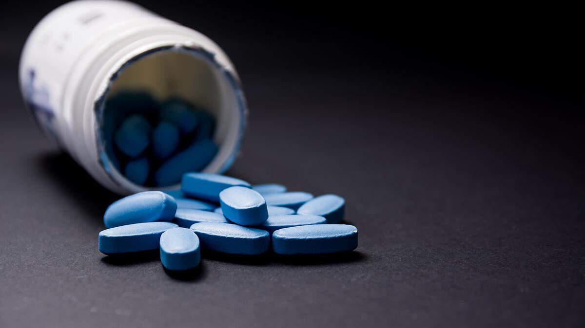 Blue pills in the foreground come out of the white plastic container in the blurred background