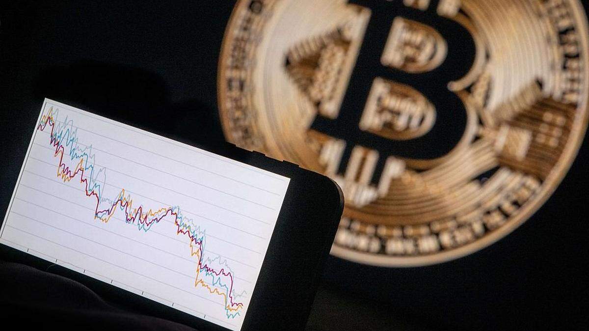This illustration photograph shows a phone with a downward trend in the stock market in front of a screen displaying a Bitcoin logo, in Mulhouse on December 5, 2025. (Photo by SEBASTIEN BOZON / AFP via Getty Images)