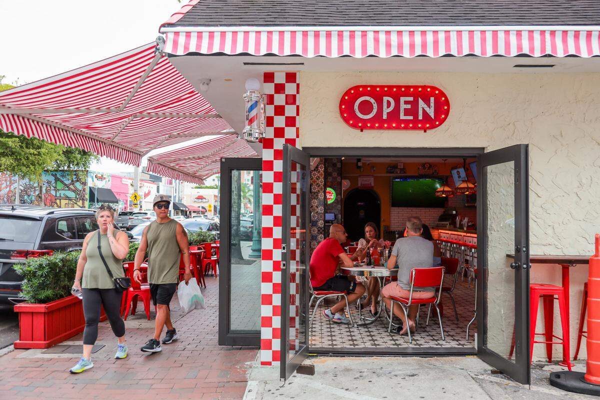 A man and a woman walk on the sidewalk as a family of tourists enjoys lunch inside the restaurant El Cuban Diner at 1620 SW Eighth St., in Miami, Fla., on Tuesday, Nov. 4, 2025.