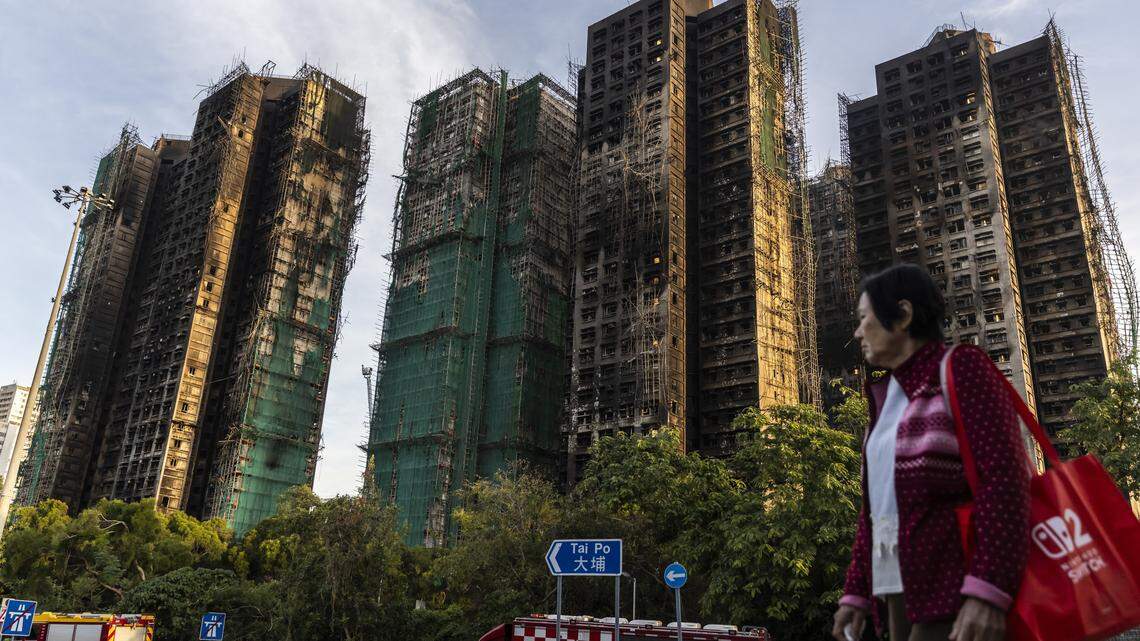 HONG KONG, CHINA - NOVEMBER 28: A woman walks past residential buildings damaged by fire at Wang Fuk Court in the Tai Po district on November 28, 2025 in Hong Kong, China. With at least 94 lives lost and rescue efforts ongoing, authorities allege gross negligence by contractors as firefighters work to fully extinguish remaining hotspots.Authorities have detained three individuals for questioning as investigators examine the cause of the blaze.