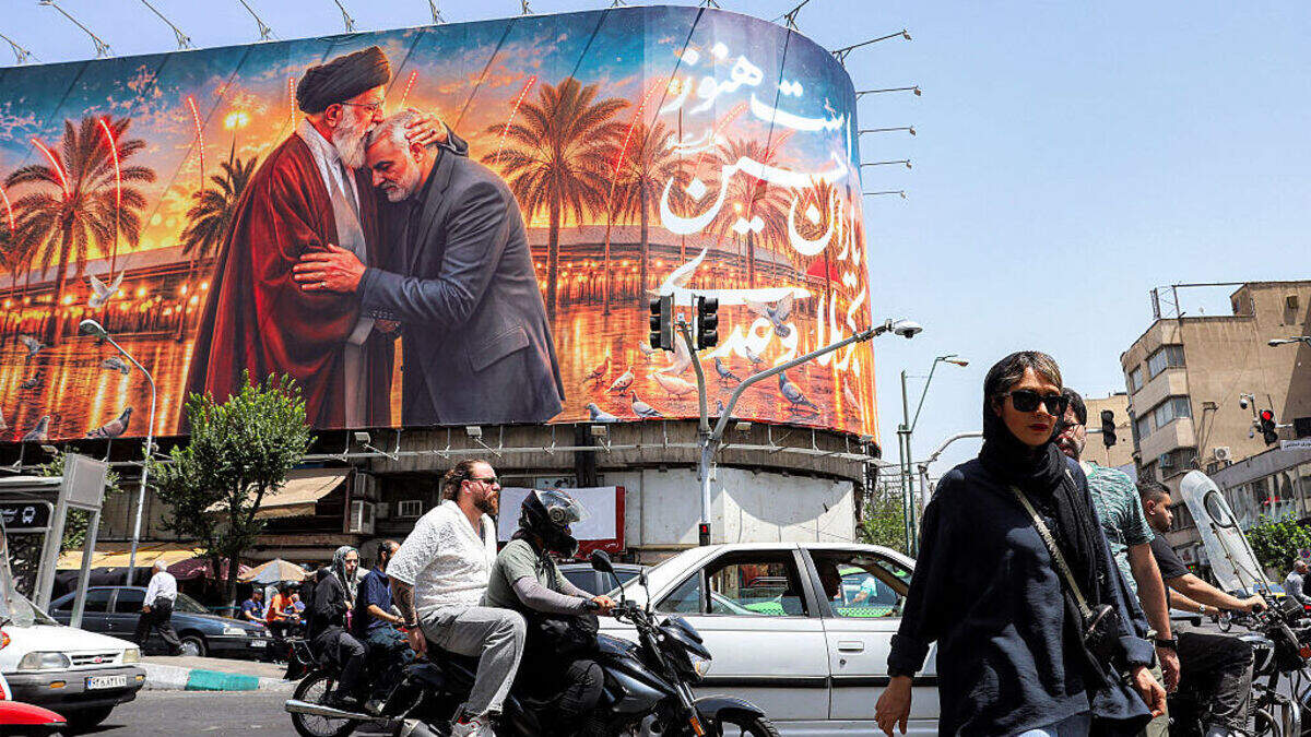 Unas personas cruzan la calle frente a una valla publicitaria —instalada en la fachada de un edificio en Teherán, el lunes 29 de junio de 2026— que muestra al fallecido líder supremo de Irán, Ali Jamenei, besando al también fallecido comandante de la Guardia Revolucionaria Islámica, Qasem Soleimani, cerca del santuario del imán Husein en la ciudad santa iraquí de Kerbala.