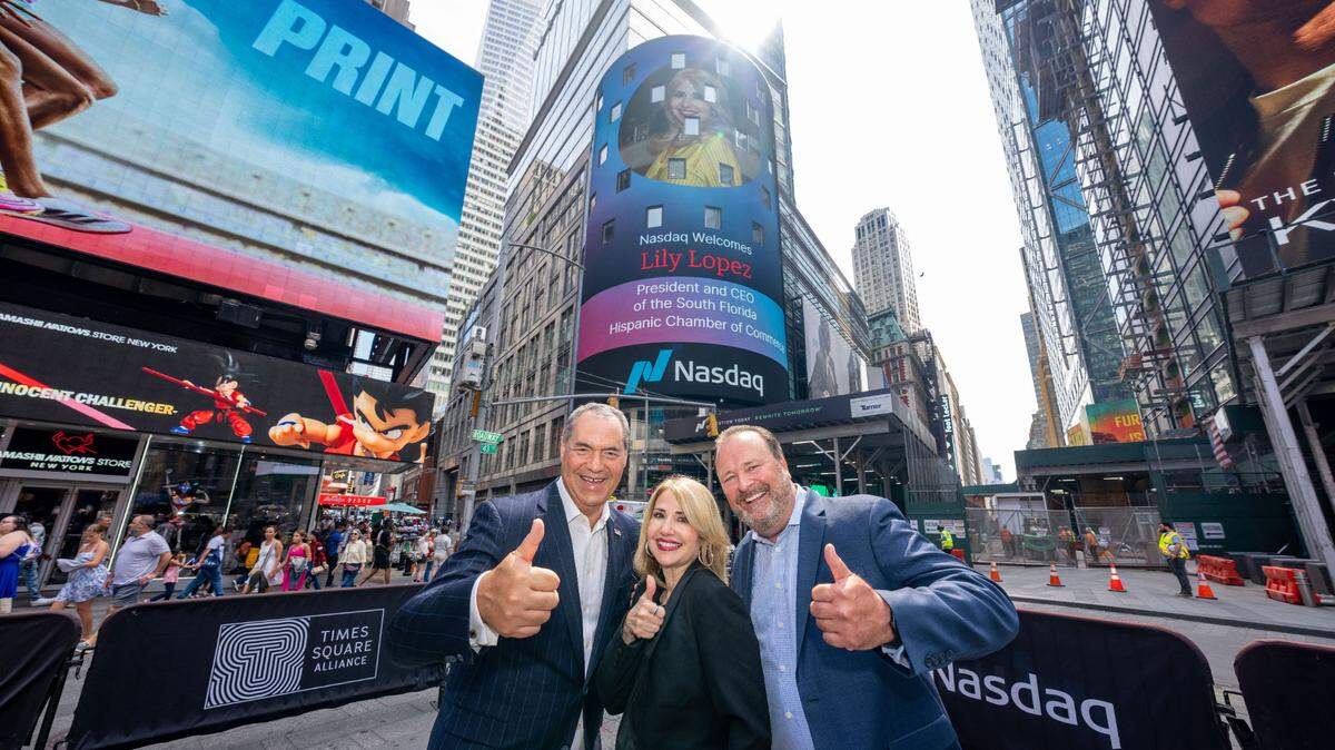 Liliam López, presidenta y fundadora de la Cámara de Comercio Hispana del Sur de la Florida, con Alex Sánchez, consejero ejecutivo del Nasdaq (der.), y David Wicks, vicepresidente del Nasdaq, frente a la pantalla de Nasdaq en Time Square, Nueva York, que ofreció un reconocimiento a López, el 25 de junio.