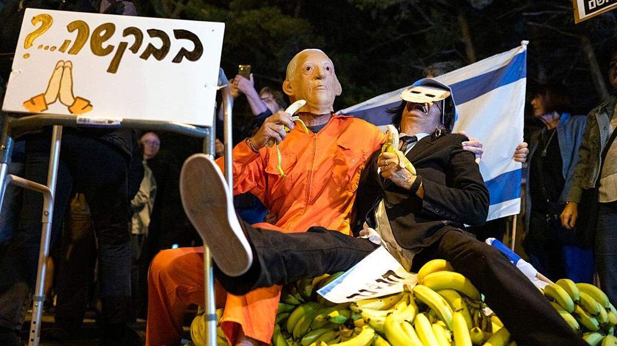A demonstrator wearing a mask depicting Israeli Prime Minister Benjamin Netanyahu sits on a pile of bananas with a man pretending to be Israeli President Isaac Herzog as people demonstrate outside Herzog's house in Tel Aviv following Netanyahu's request for a presidential pardon on November 30, 2025. Israeli Prime Minister Benjamin Netanyahu, on trial facing corruption charges, announced on November 30 he had submitted a pardon request, saying the long-running cases were tearing the country apart. US President Donald Trump wrote to Israeli President Isaac Herzog earlier this month, asking him to pardon Netanyahu, who has repeatedly denied wrongdoing in the proceedings. (Photo by Maya Levin / AFP via Getty Images)