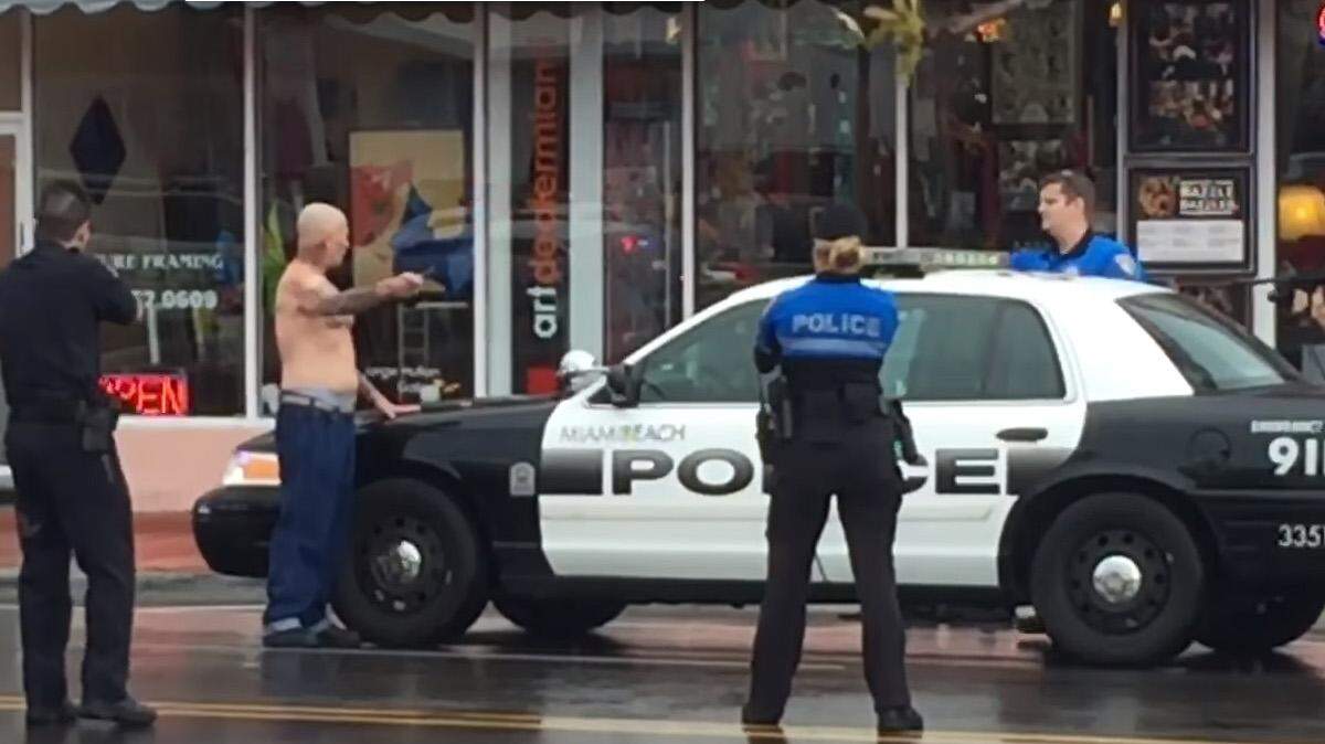 En el video, un hombre calvo, sin camisa y sosteniendo una navaja de barbero en la mano izquierda da un paso hacia dos agentes de la policía de Miami Beach, que apuntan sus armas hacia él. 