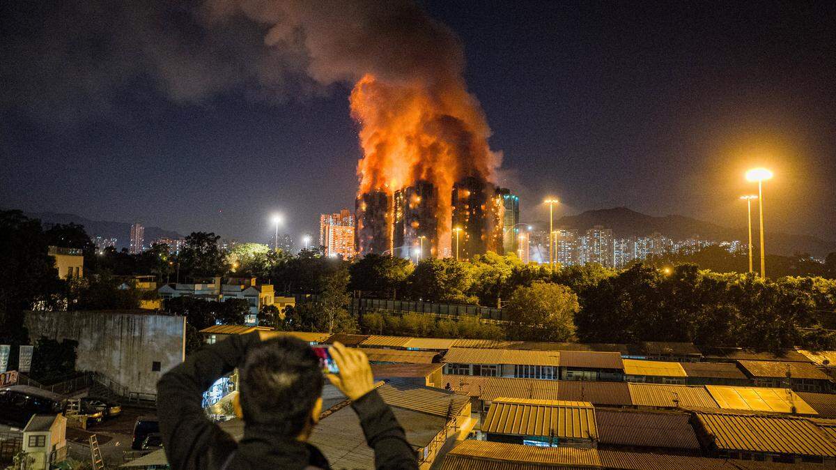 An onlooker takes photos as a major fire engulfs several apartment blocks at the Wang Fuk Court residential estate (background) in Hong Kong's Tai Po district on November 26, 2025. At least four people were killed when a fire engulfed several high-rise blocks in a Hong Kong residential estate on November 26, the government said, with media reporting that some residents were trapped inside. (Photo by AFP via Getty Images)