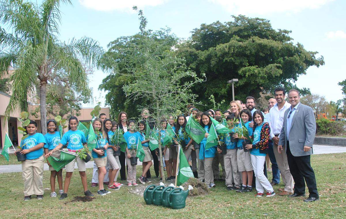 Artista Xavier Cortada y otros adultos posan con una clase de niños de Sweetwater Elementary, todos quienes tienen una bandera verde símbolo del proyecto "Native Flags" y un árbol nativo a la Florida. Los niños tienen camiseta de uniforme turquesa. Todos están afuera, y hay arboles detrás ellos.