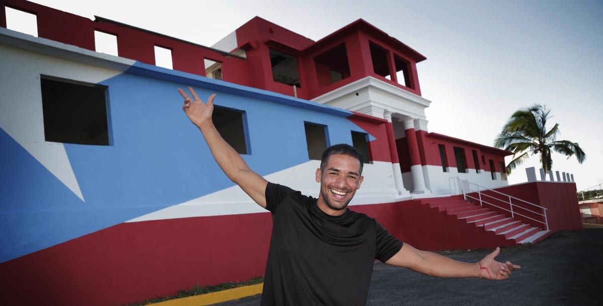 El artista Héctor Collazo Hernández frente a la bandera número 60 del proyecto 78 pueblos y una bandera, pintada en un hospital abandonado de Barceloneta, Puerto Rico.