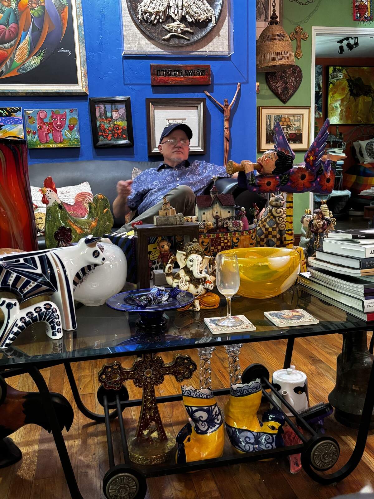 Julio Taquechel en la sala de su casa rodeado de mesas repletas de libros, cerámicas, jarrones, cajas pequeñas y esculturas.