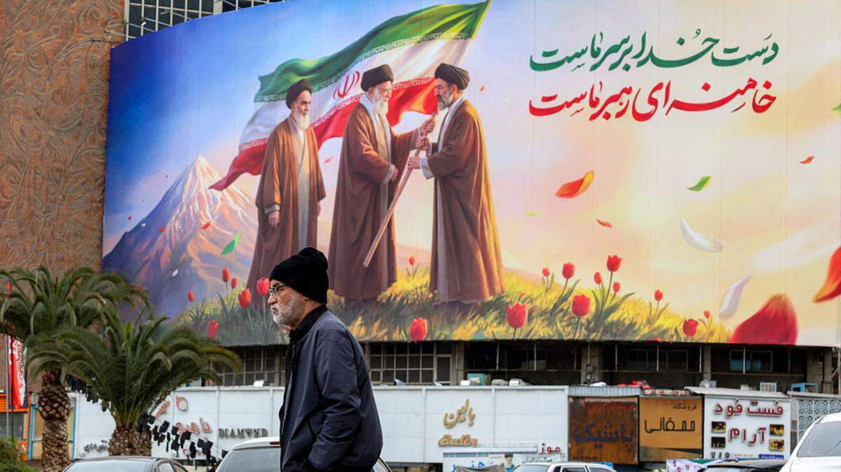 Un hombre camina cerca de una pancarta exhibida en la plaza Valiasr en el centro de Teherán, el martes 10 de marzo de 2026, que representa al difunto líder supremo de Irán, el ayatolá Ruhollah Jomeini (izq.), observando cómo su sucesor, el difunto ayatolá Alí Jamenei (centro), entrega una bandera nacional a su hijo y nuevo líder supremo, Mojtaba Jamenei (der.).