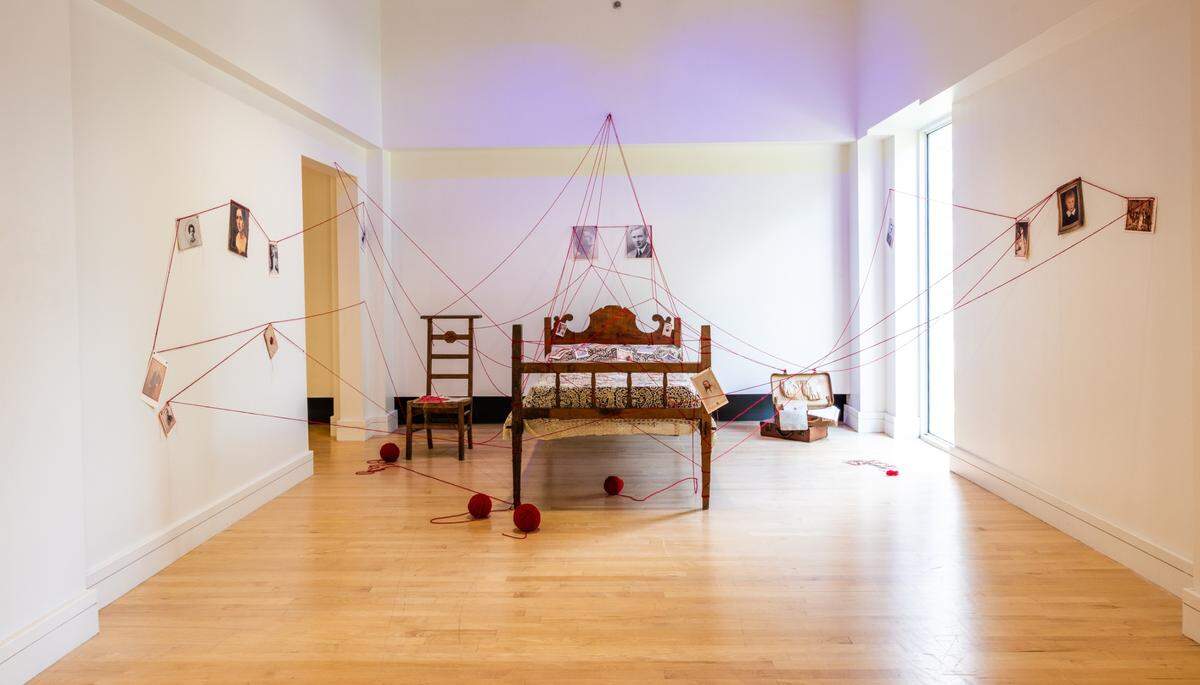 'Ancestral Lines-Inherited Ties', 2025, de Marine Fonteyne. Antigua cama de familia, cobertor con encaje de Brujas (Bélgica), fotografías familiares bordadas, lazo rojo y otros objetos personales.