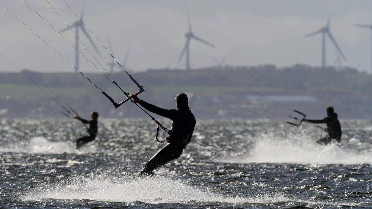Surfistas disfrutan de la brisa marina, con molinos de energía eólica en el fondo, en una playa de la costa oeste de la isla de Fehmarn, Alemania, el 15 de octubre de 2024.